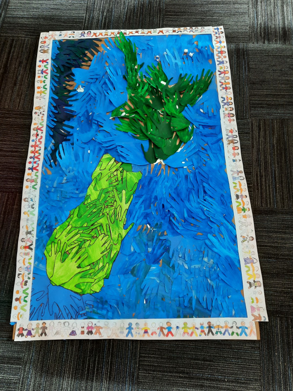 Artwork created by students at St Matthews Primary School in Hastings for the theme “Together We Belong”. Hundreds of painted handprints are arranged to form New Zealand, surrounded by blue handprints representing the Pacific Ocean. Around the artwork are paper cut-out figures coloured with national flags and traditional clothing from more than 15 countries represented in the school.