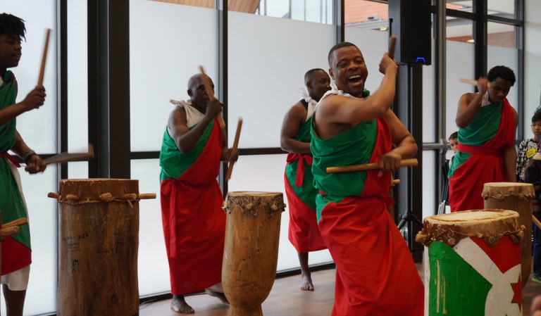 Five Burundi drummers perform energetically while smiling and enjoying performing for the crowd at the INZ pōwhiri for refugees