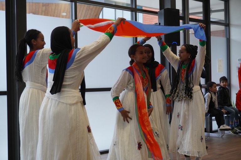 Five Eritrean performers dance under their flag in long flowing white dresses smiling and enjoying performing at the INZ pōwhiri for refugees